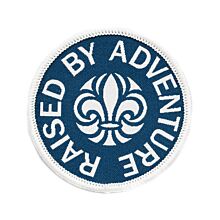 Runt märke med vit langetterad kant och blå bakgrund med scoutsymbolen på och runt den texten Raised By Adventure