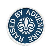 Runt märke med vit langetterad kant och blå bakgrund med scoutsymbolen på och runt den texten Raised By Adventure