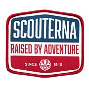 Märke med röd langetterad kant, vit bakgrund och blå detalj med texten Scouterna Raised By Adventure since 1910 och scoutsymbolen på